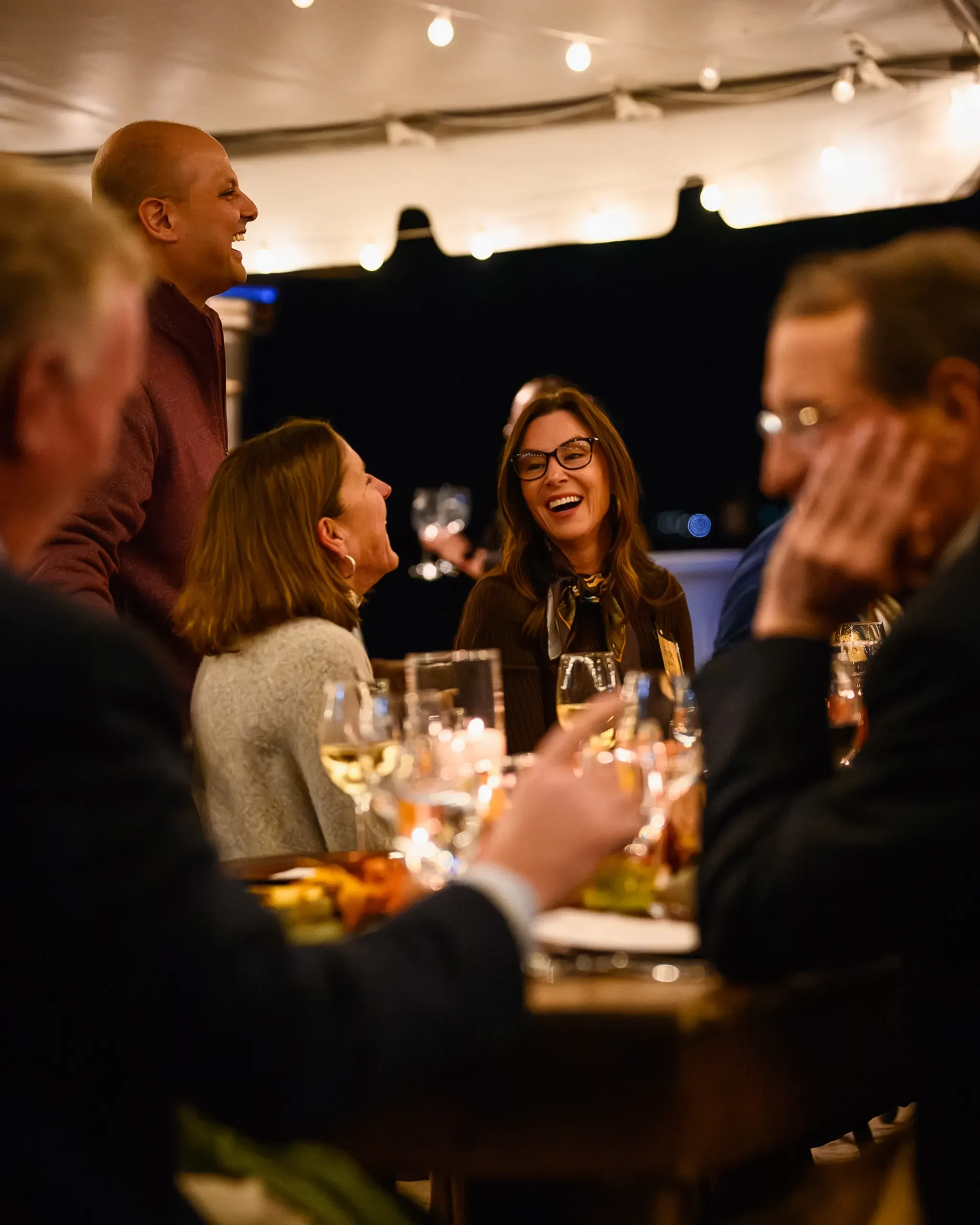 Guests laughing around a candlelit dinner table under string lights at the Bolles Harvest Dinner