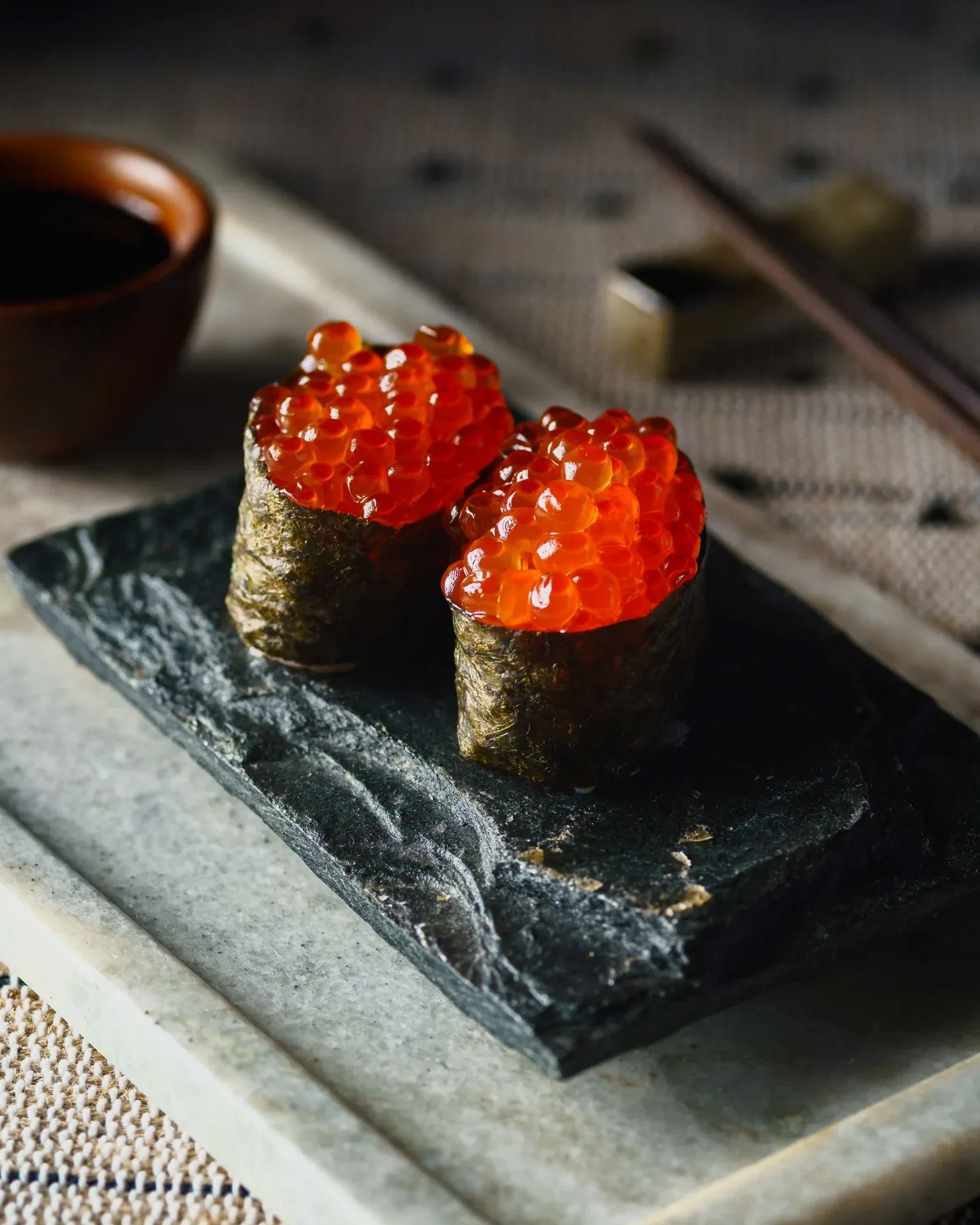 Two ikura gunkan maki on black slate with soy sauce and chopsticks