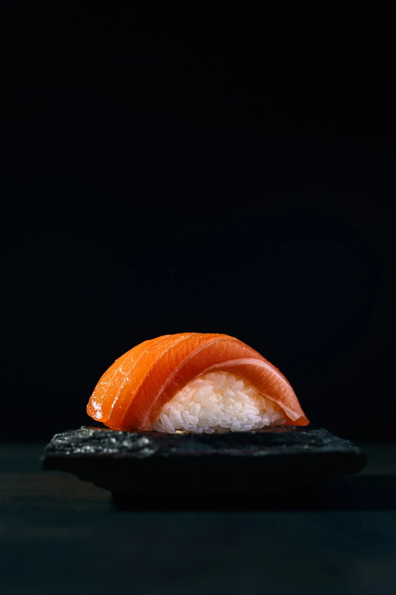 Single salmon nigiri on dark stone with black background